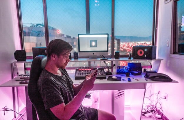 A gamer in a brightly lit room surrounded by multiple monitors, a professional microphone, and a high-quality camera. The gamer, wearing a headset, is intensely focused on the game displayed on the central monitor, with live chat and game statistics visible on the side screens. The setup indicates a professional streaming environment.